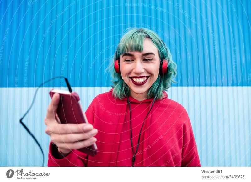 Porträt einer lachenden jungen Frau mit blau gefärbtem Haar und Kopfhörern, die ein Selfie mit einem Smartphone macht blaue blauer blaues faerben färben