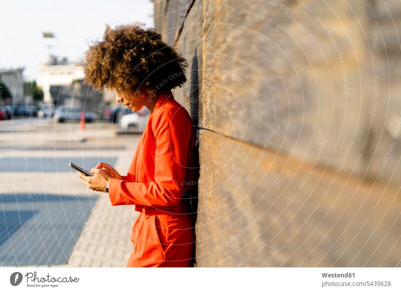 Junge Frau in modischem roten Hosenanzug lehnt an der Wand und benutzt ein Mobiltelefon Leute Menschen People Person Personen gemischtrassig 1 Ein ein Mensch