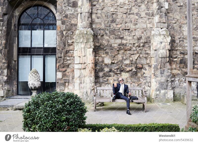 UK, London, älterer Geschäftsmann sitzt auf einer Bank in einem Innenhof und entspannt sich, während er mit Kopfhörern Musik hört Entspannung relaxen entspannen