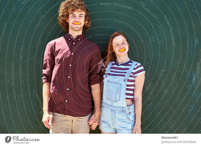 Junges Paar steht vor einer grünen Wand mit orangefarbenen Scheiben im Mund stehen stehend Stück Orange Apfelsinen Orangen Wände Waende Pärchen Paare