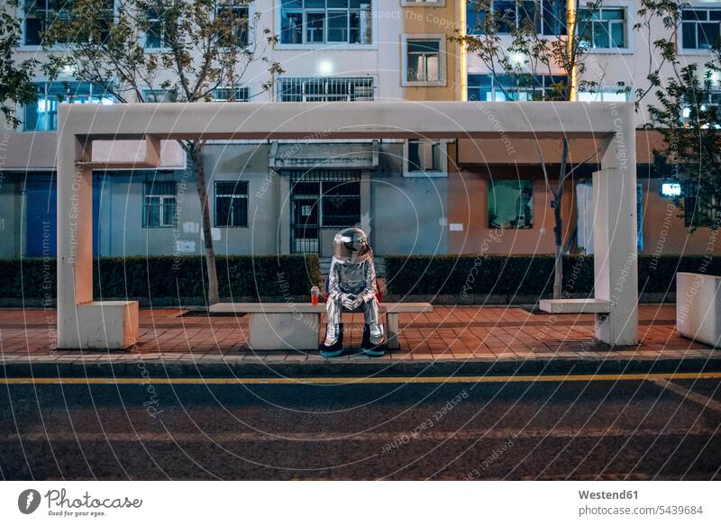Spaceman sitzt auf Bank an einer Bushaltestelle in der Nacht mit Softdrink Haltestelle Haltestellen Bushaltestellen Raumfahrer Weltraumfahrer nachts Astronaut