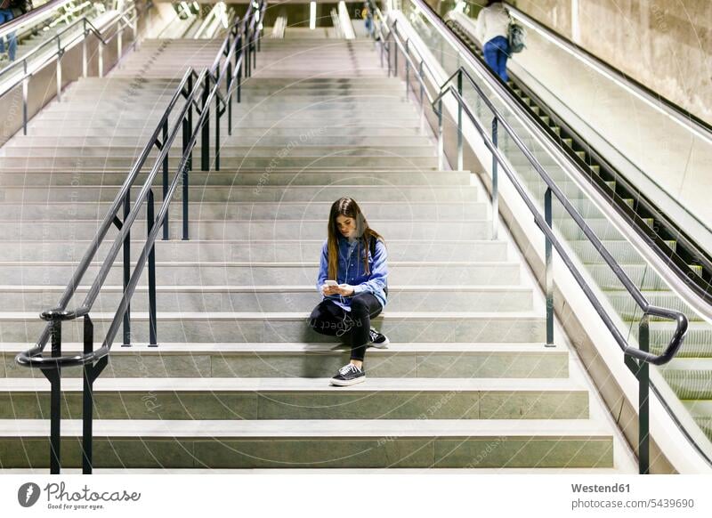Junge Frau sitzt auf der Treppe und benutzt Handy und Kopfhörer Smartphone iPhone Smartphones benutzen weiblich Frauen sitzen sitzend Ohrhörer Mobiltelefon