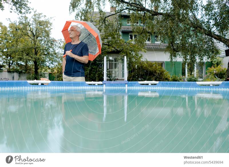 Älterer Mann steht im Sommer regen im Garten am Schwimmbad hält Regenschirm Gaerten Gärten stehend Jahreszeiten sommerlich Sommerzeit Schirme Regenschirme Leute