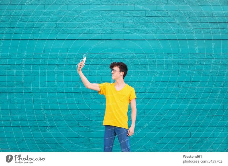 Junger Mann macht ein Selfie mit Smartphone vor einer blauen Backsteinmauer Männer männlich Selfies Erwachsener erwachsen Mensch Menschen Leute People Personen