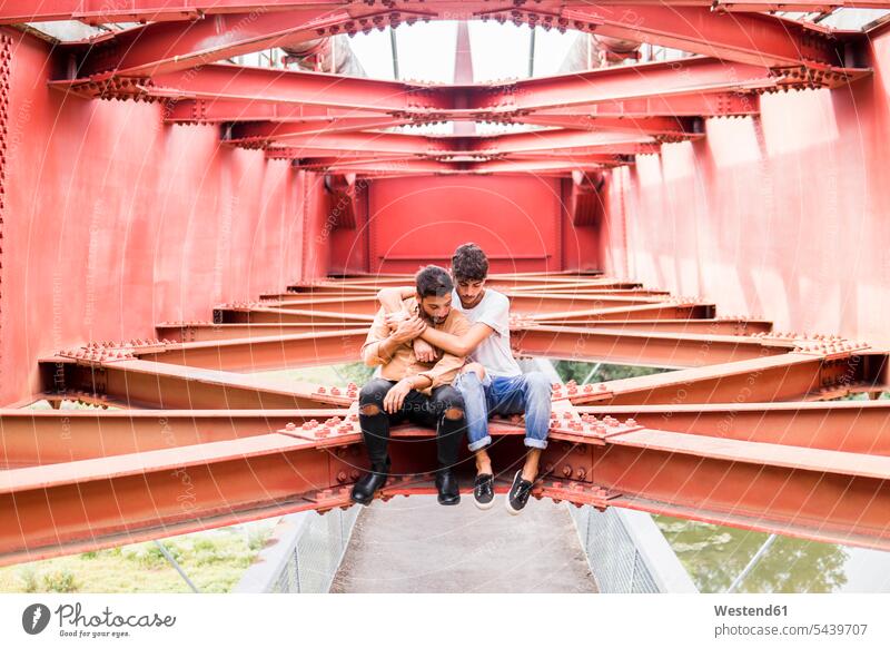 Junges schwules Liebespaar sitzt auf Stahlträger einer Fußgängerbrücke und schaut nach unten Paar Pärchen Paare Partnerschaft Fußgängerbrücken