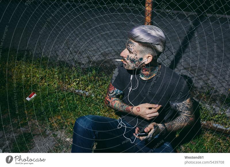 Tätowierter junger Mann mit Ohrstöpseln und Smartphone raucht eine Zigarette am Maschendrahtzaun tätowiert Tattoo Tätowierungen Tatoos Taetowierung Tattoos