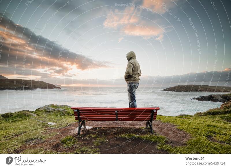 Spanien, Ferrol, Mann mit Kapuzenjacke steht auf einer Bank an der Küste und schaut in die Ferne Kueste Kuesten Küsten Außenaufnahme draußen im Freien