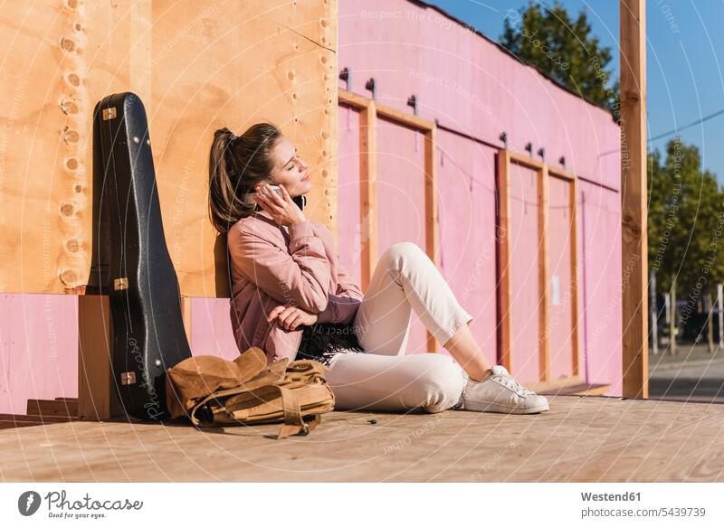 Lächelnde junge Frau sitzt auf einer Plattform neben einem Gitarrenkoffer und hört Musik sitzen sitzend weiblich Frauen zuhören zuhörend lächeln hoeren