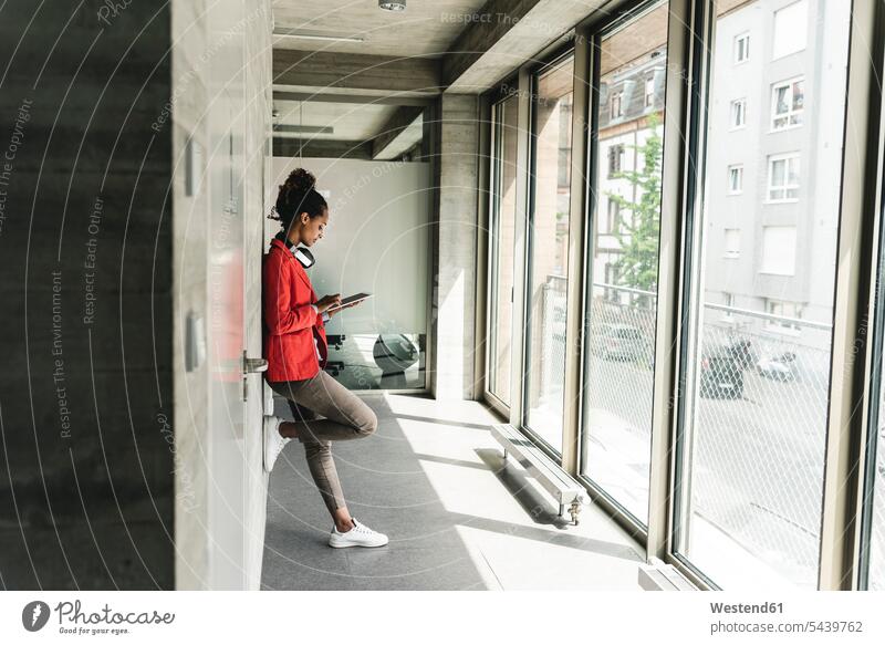 Junge Frau mit Kopfhörern steht im Flur und benutzt ein digitales Tablett Kreative Kreativer Kreativer Beruf Vernetzung Networking stehen stehend Fenster