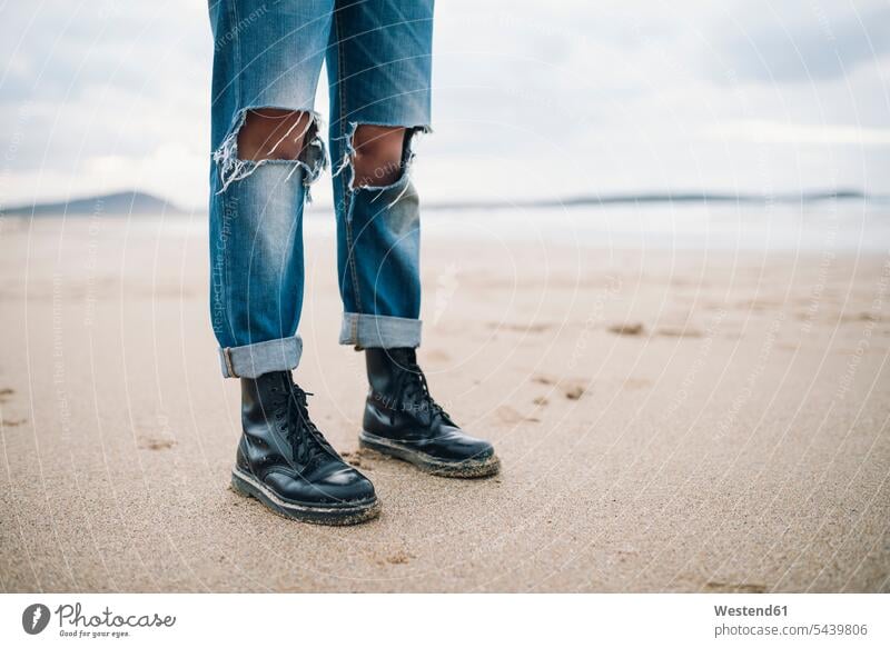 Frau mit Stiefeln und zerrissenen Jeans am Strand, Teilansicht Beach Straende Strände Beaches weiblich Frauen zerfetzt gerissen Jeanshose stehen stehend steht