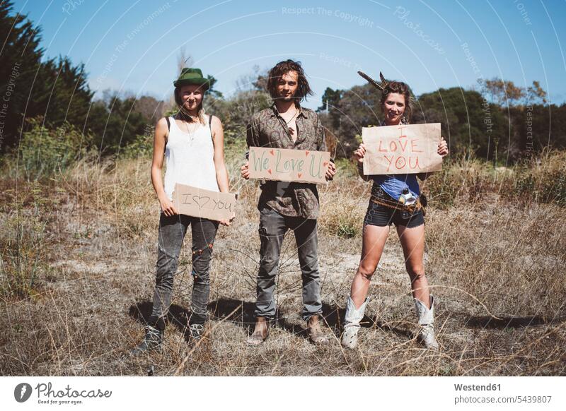 Drei Hippies halten "Ich liebe dich"-Schilder in der Natur Öde Einöde lächeln sympathisch nett Freizeitkleidung casual Freizeitbekleidung Tag am Tag