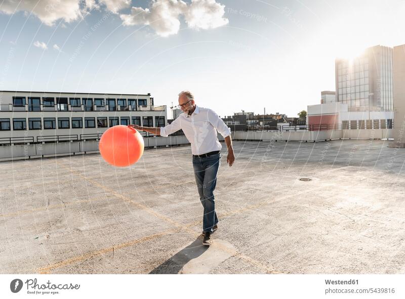 Erwachsener Mann spielt mit orangefarbenem Fitnessball auf dem Dach eines Hochhauses Businessmann Textfreiraum Himmel modern Wolke Wolken Tag am Tag
