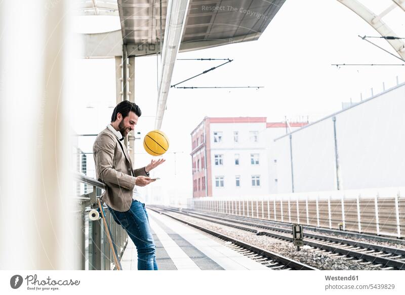 Lächelnder junger Mann mit Handy und Basketball auf dem Bahnsteig Mobiltelefon Handies Handys Mobiltelefone lächeln Bahnhof Bahnhöfe Bahnhoefe Zugbahnsteig