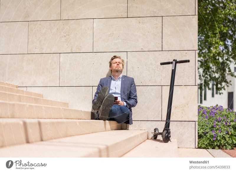 Geschäftsmann mit E-Scooter und Smartphone sitzt entspannt auf Stufen geschäftlich Geschäftsleben Geschäftswelt Geschäftsperson Geschäftspersonen Businessmann