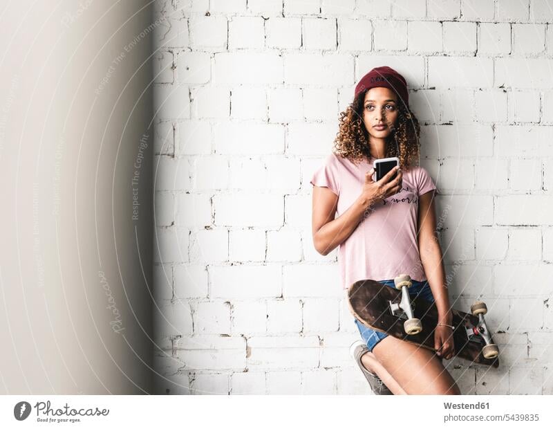 Porträt einer jungen Frau mit Skateboard, an die Wand gelehnt Mauer Mauern junge Frau junge Frauen anlehnen angelehnt lehnend Wollmütze Wollmützen Strickmütze