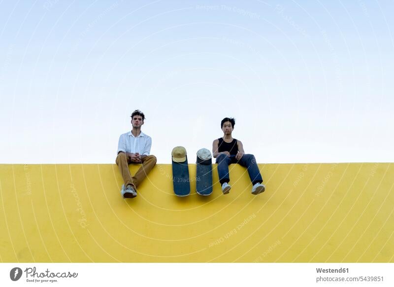 Zwei junge Männer mit Skateboards sitzen auf einer hohen gelben Wand Freunde Mann männlich Mauer Mauern hoch gelber gelbes ausruhen Rast Erholung erholen