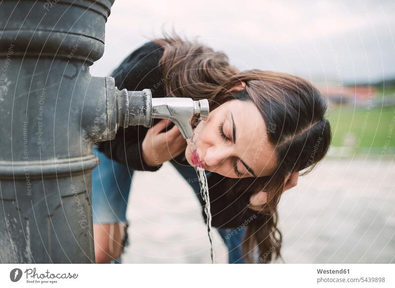 Junge Frau trinkt Wasser aus einem Brunnen trinken weiblich Frauen Erwachsener erwachsen Mensch Menschen Leute People Personen stehen stehend steht Wasserhahn