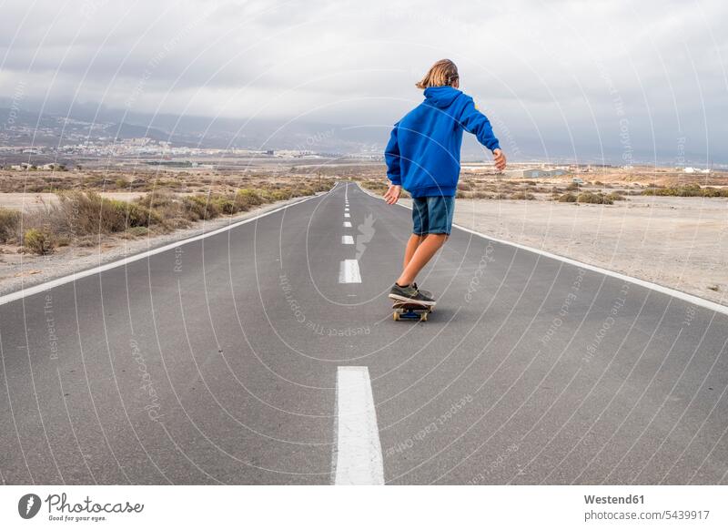 Spanien, Teneriffa, Rückenansicht eines Jungen, der auf einer leeren Landstraße Skateboard fährt Straße Strassen Straßen Buben Knabe Knaben männlich Rollbretter