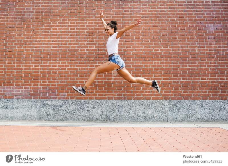 Überschwängliche junge Frau springt vor eine Ziegelmauer Spaß Spass Späße spassig Spässe spaßig Ziegelwand Backsteinmauer glücklich Glück glücklich sein
