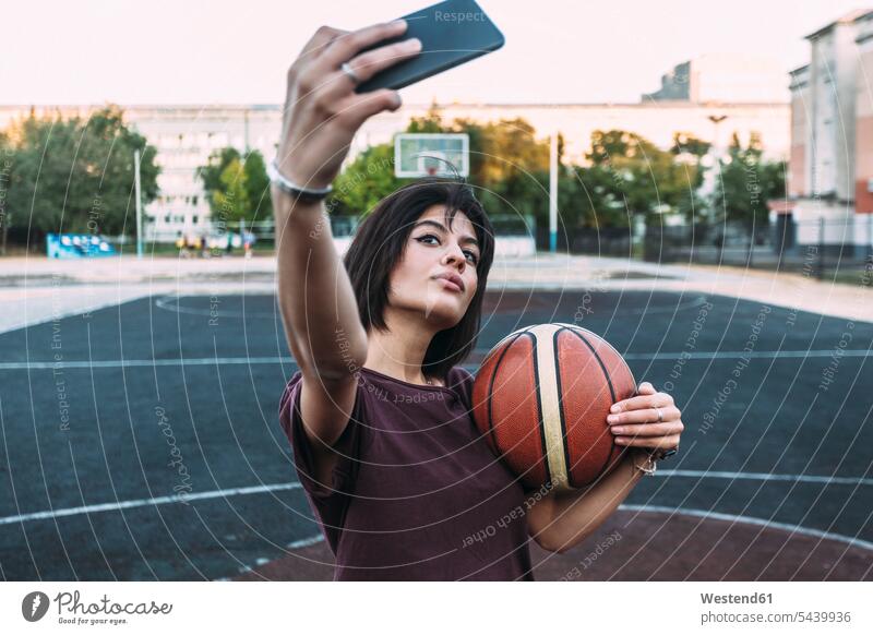Junge Frau mit Basketball macht ein Selfie auf einem Platz im Freien weiblich Frauen Selfies Basketbaelle Basketbälle Sportplatz Sportplaetze Sportplätze