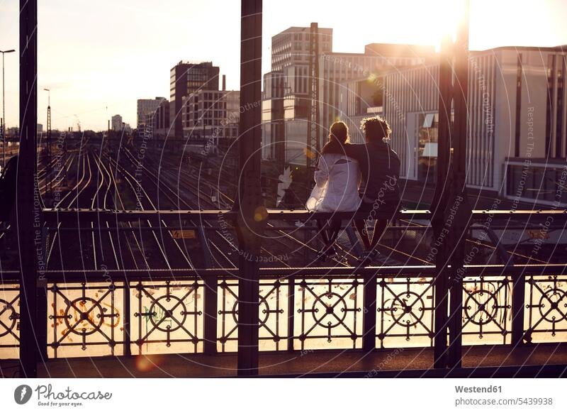 Deutschland, München, Junges Paar sitzt auf der Brücke und genießt den Sonnenuntergang Sonnenuntergänge umarmen Umarmung Umarmungen Arm umlegen Zukunft sitzen
