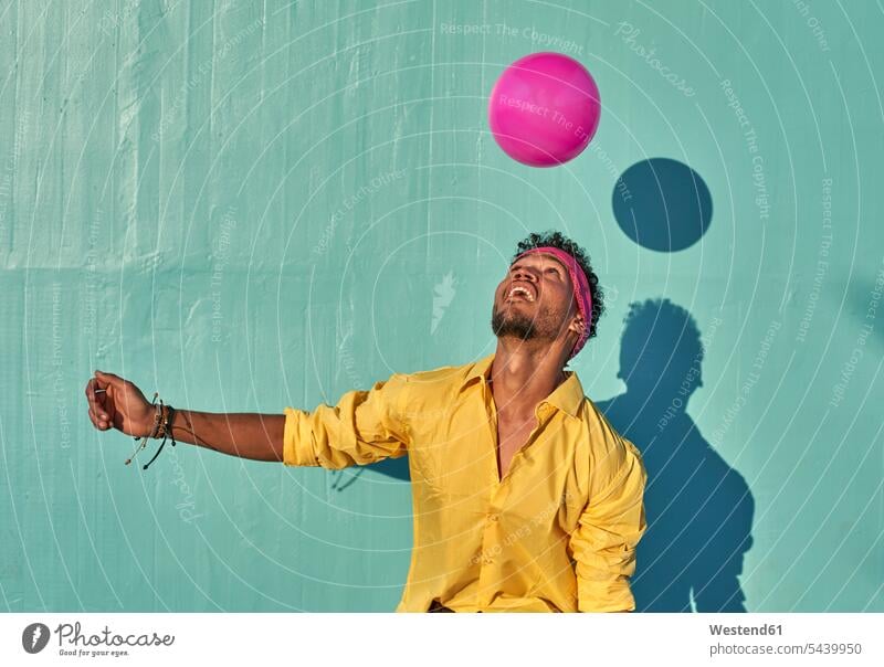 Junger schwarzer Mann spielt mit einem rosa Ball vor einer blauen Wand Hemden Bälle freuen Glück glücklich sein glücklichsein gefühlvoll Emotionen Empfindung