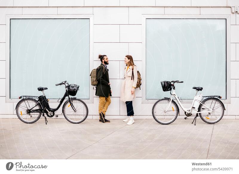 Mann und Frau mit E-Bikes stehen vor einem Gebäude und unterhalten sich stehend steht eBikes Elektrofahrrad Elektrorad Paar Pärchen Paare Partnerschaft sprechen
