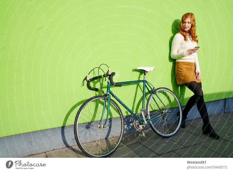 Junge Frau mit einem Handy, das gegen eine grüne Wand neben dem Fahrrad lehnt Bikes Fahrräder Räder Rad Mobiltelefon Handies Handys Mobiltelefone weiblich