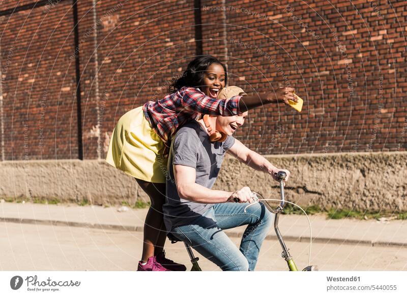 Junges Paar fährt Fahrrad auf der Straße, Frau steht auf dem Gepäckträger und macht Selfies radfahren fahrradfahren radeln Handy Mobiltelefon Handies Handys