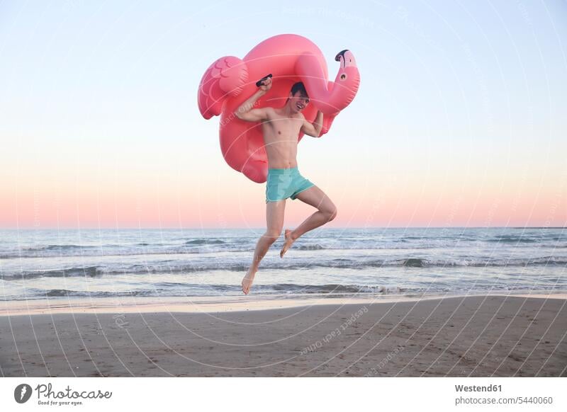 Lachender junger Mann springt am Strand mit aufblasbarem rosa Flamingo in die Luft Beach Straende Strände Beaches springen hüpfen Männer männlich Sprung