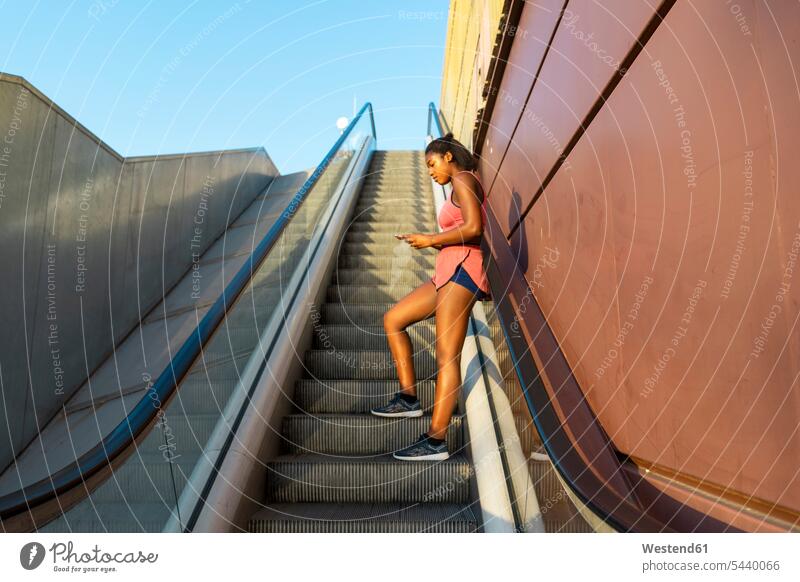 Junge sportliche Frau auf Rolltreppe mit Smartphone benutzen benützen Fahrtreppe Sportlerin Sportlerinnen iPhone Smartphones Workout weiblich Frauen Handy