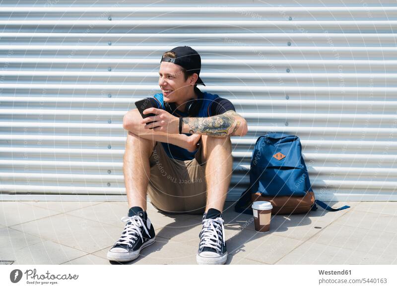 Lächelnder junger Mann mit Smartphone, Rucksack und Kaffee zum Sitzen auf dem Boden warten Männer männlich Erwachsener erwachsen Mensch Menschen Leute People