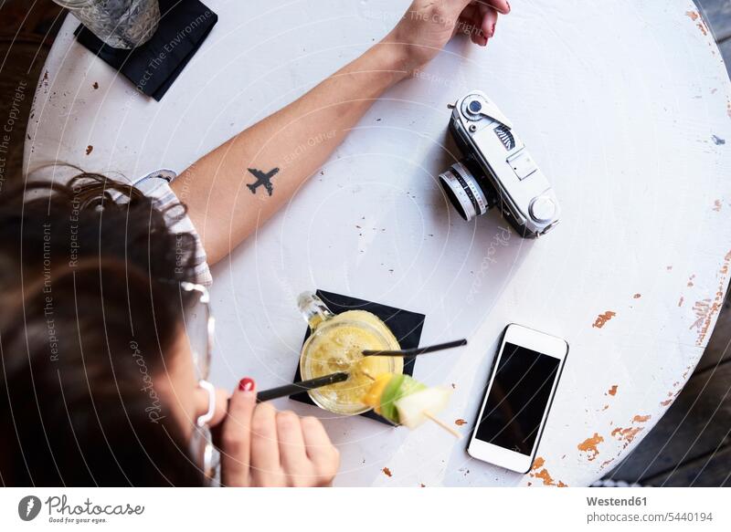 Junge Frau mit Flugzeugtätowierung auf dem Unterarm sitzt in einem Café und trinkt Ananas-Smoothie Cafe Kaffeehaus Bistro Cafes Kaffeehäuser Smoothies