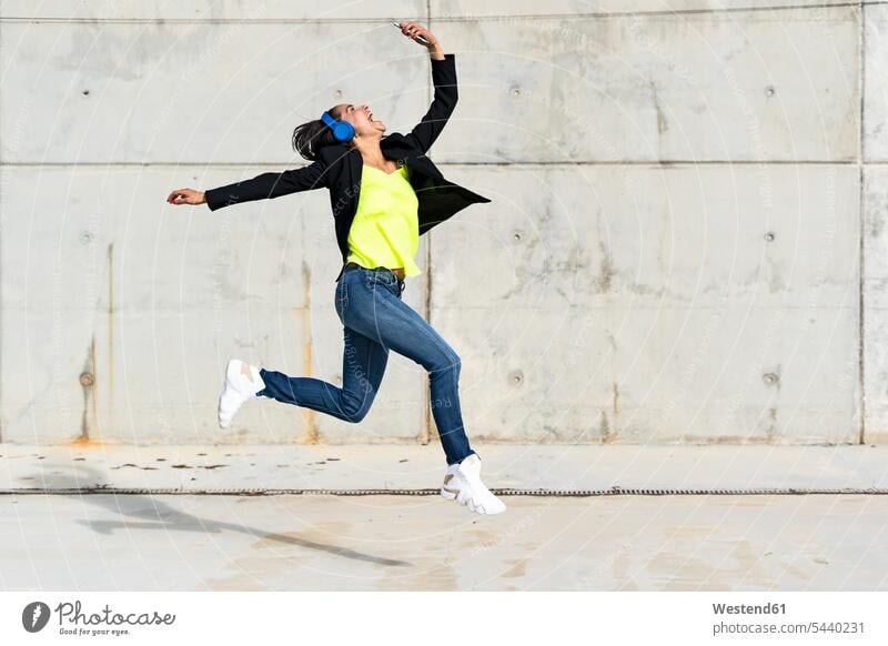 Frau mit blauen Kopfhörern hört Musik, springt in die Luft und macht ein Selfie Spanien Drahtlose Technologie drahtlose Verbindung Drahtlose Kommunikation