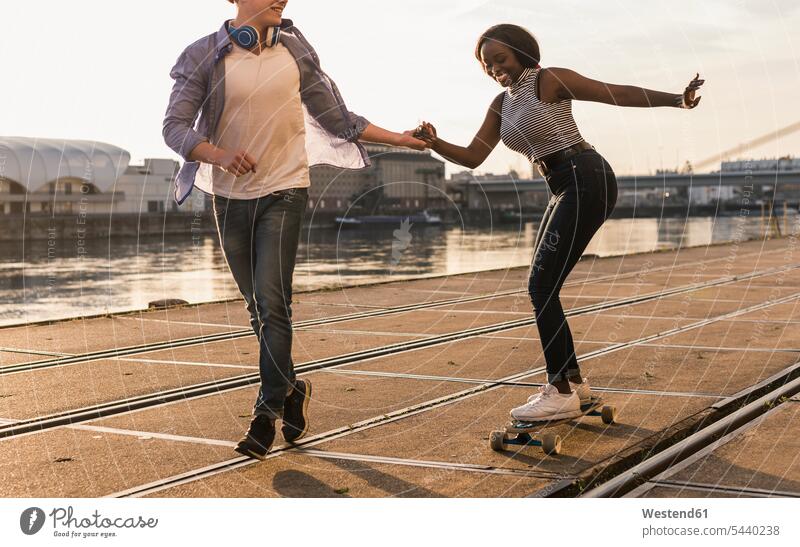 Junges Paar beim Skateboarden am Flussufer multikulturell ziehen Hände halten fröhlich jung aktiv Freizeitsport Freizeitaktivität Hand in Hand Freude Aktivität