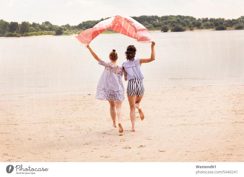 Rückenansicht von zwei Freunden, die nebeneinander am Strand laufen und ein Tuch über den Kopf halten Beach Straende Strände Beaches Freundinnen rennen
