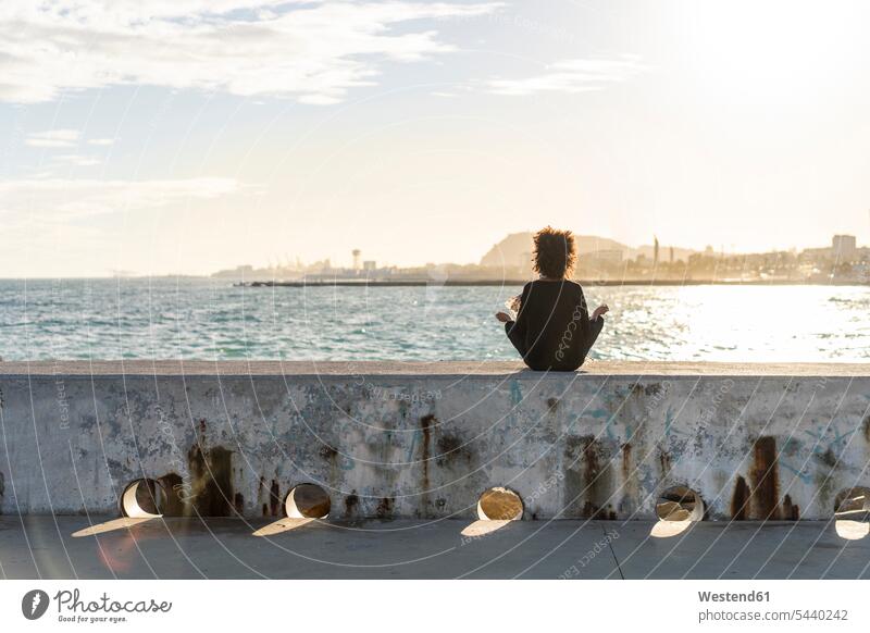 Spanien, Barcelona, Rückenansicht einer Frau, die auf einer Mauer vor dem Meer sitzt und meditiert sitzen sitzend meditieren Meere weiblich Frauen Mauern