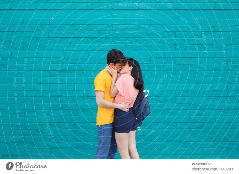 Junges Paar küsst sich vor blauer Ziegelmauer küssen Küsse Kuss Liebespaar Liebespaare Pärchen Paare Partnerschaft Mensch Menschen Leute People Personen stehen