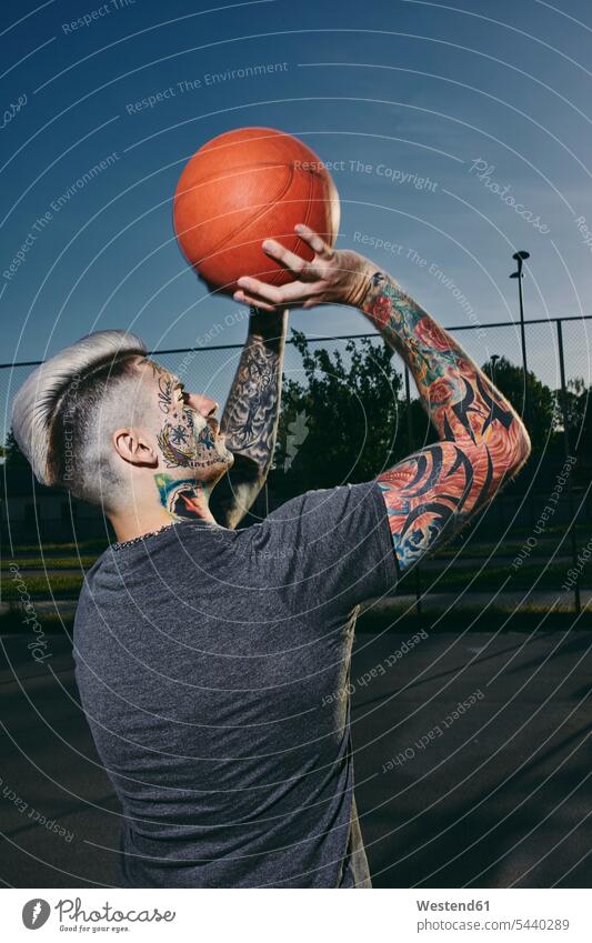 Tätowierter junger Mann wirft Basketball im Freien tätowiert Tattoo Tätowierungen Tatoos Taetowierung Tattoos Taetowierungen Sportplatz Sportplaetze Sportplätze