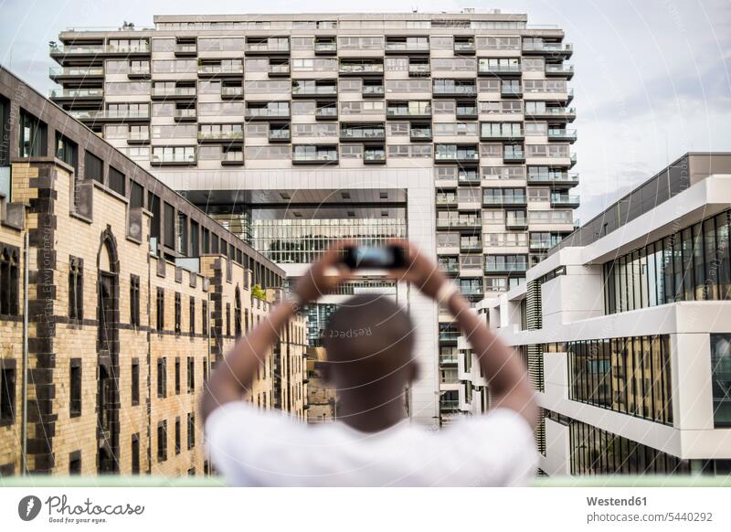 Deutschland, Köln, junger Mann beim Fotografieren mit Smartphone Schwarzer Farbige Farbiger dunkelhäutig Afrikanische Abstammung Gebäude Gebaeude Rheinauhafen