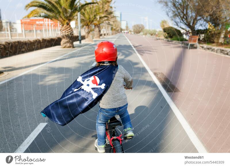 Spanien, Barcelona, Rückenansicht eines kleinen Jungen mit einem Piratenumhang, der auf einer leeren Straße Fahrrad fährt Kindheit Fahrradtour Fahrradausflug