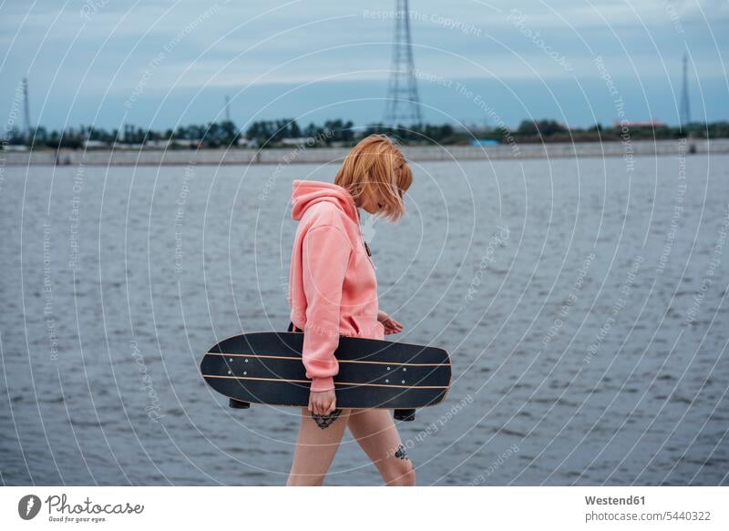 Junge Frau hält Carver Skateboard zu Fuß am Flussufer Rollbretter Skateboards Fluesse Fluß Flüsse halten weiblich Frauen gehen gehend geht Ufer Gewässer Wasser