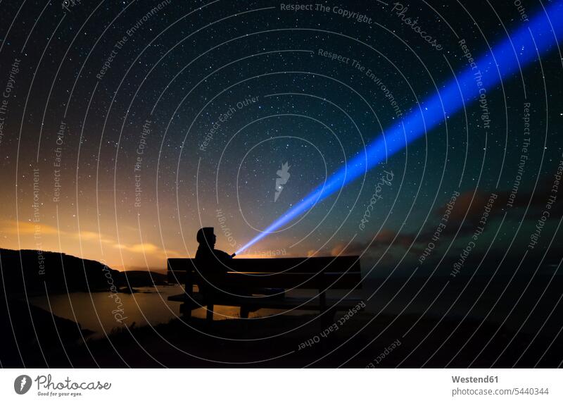 Spanien, Ortigueira, Loiba, Silhouette eines auf Bank sitzenden Mannes unter Sternenhimmel mit blauem Strahl Bänke Sitzbank Sitzbänke Ruhe Beschaulichkeit ruhig