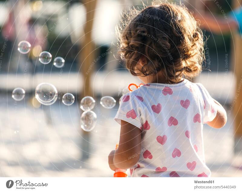 Rückenansicht eines kleinen Mädchens, das auf dem Spielplatz Seifenblasen macht Europäer Kaukasier kaukasisch Europäisch Tag tagsueber Tageslichtaufnahme