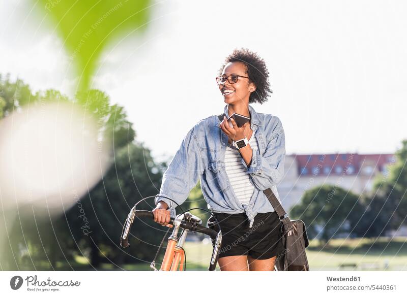 Lächelnde junge Frau mit Handy und Fahrrad im Park Bikes Fahrräder Räder Rad Mobiltelefon Handies Handys Mobiltelefone Parkanlagen Parks weiblich Frauen lächeln