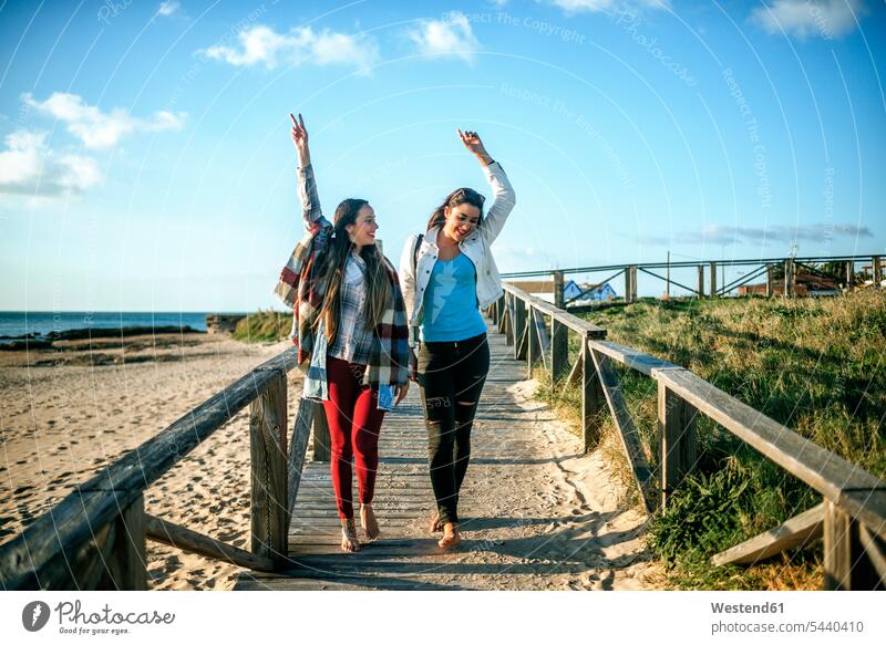 Zwei glückliche junge Frauen gehen barfuss auf der Promenade Freundinnen Holzsteg Holzweg Fußweg Bohlenweg Freunde Freundschaft Kameradschaft Steg Strand Beach