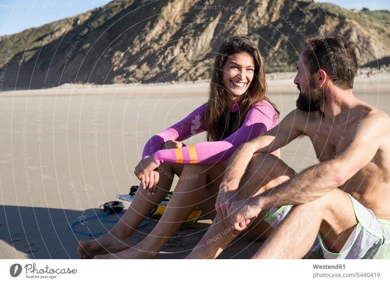 Lächelndes Paar mit Surfbrett am Strand sitzend Beach Straende Strände Beaches sitzt Surfbretter surfboard surfboards Pärchen Paare Partnerschaft lächeln Surfer