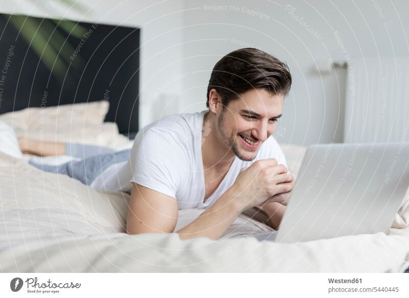 Lächelnder Mann liegt mit Laptop auf dem Bett Betten Notebook Laptops Notebooks lächeln Männer männlich Computer Rechner Erwachsener erwachsen Mensch Menschen