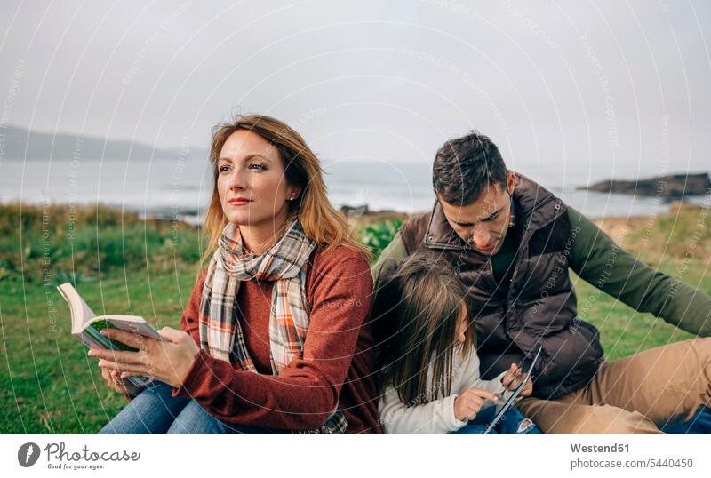Entspannte Frau mit Buch, die zur Seite schaut, während Mädchen und Mann im Freien Tabletten benutzen Familie Familien Küste Kueste Kuesten Küsten Mensch