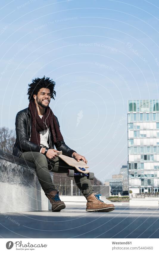 Lächelnder Mann mit Longboard sitzt im Skatepark und hört Musik auf seinem Smartphone Handy Mobiltelefon Handies Handys Mobiltelefone Männer männlich